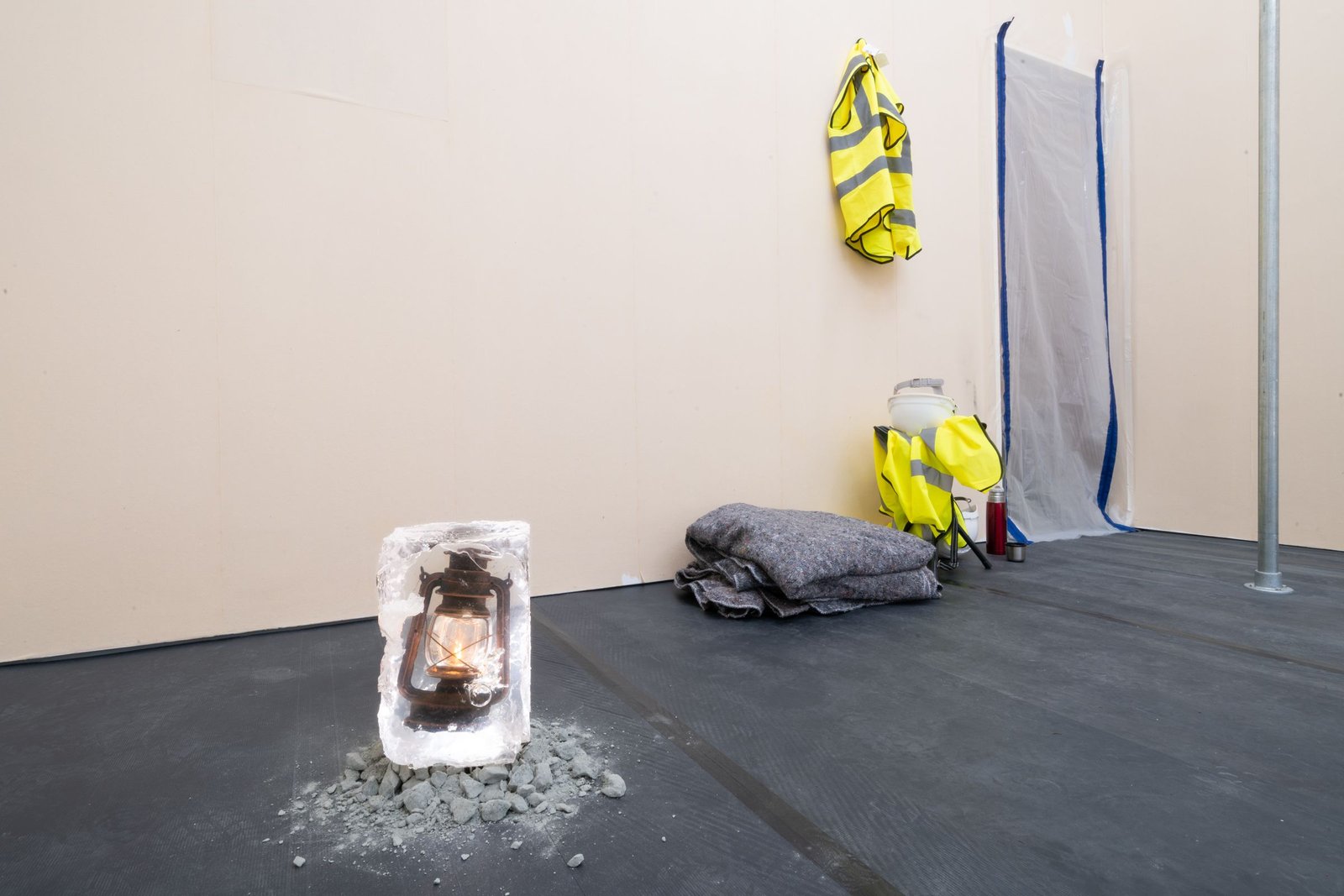 A lantern encased in ice sits on gravel. Nearby, a folded blanket, a yellow safety vest, and a plastic container are placed against a light-colored wall.